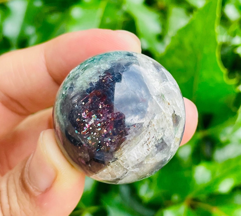 May include: A clear, spherical crystal ball with internal inclusions of dark red, green, and iridescent glitter. The sphere is held in a hand against a backdrop of green foliage. The crystal ball has a polished, reflective surface.