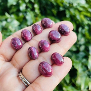 Puede incluir: Una colección de nueve piedras preciosas de rubí de forma ovalada, talladas en cabujón. Las piedras son de un color rojo intenso y rico y tienen un acabado liso y pulido.