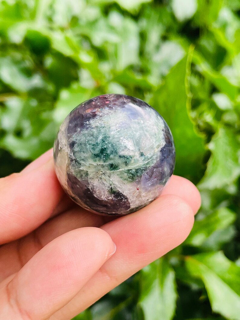 May include: A polished fluorite sphere, showcasing a blend of purple, green, and clear crystalline structures. The gemstone ball is held in a hand, with a blurred green foliage background. The sphere's surface reflects light, highlighting its natural patterns.