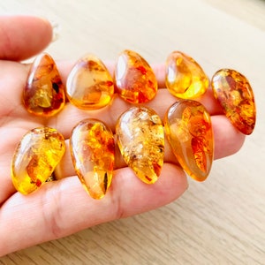 Op de afbeelding: Een verzameling van tien amber druppel hangers, elk met een uniek patroon en kleurvariatie. De hangers zijn een warme, goudgele kleur en hebben een gladde, gepolijste afwerking.