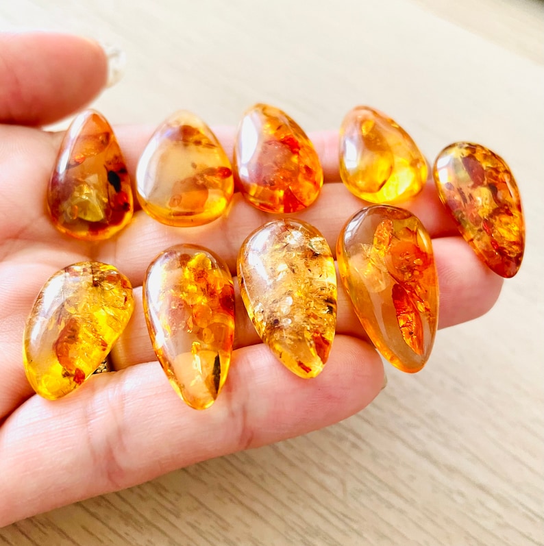 Op de afbeelding: Een verzameling van tien amber druppel hangers, elk met een uniek patroon en kleurvariatie. De hangers zijn een warme, goudgele kleur en hebben een gladde, gepolijste afwerking.