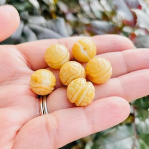 Könnte beinhalten: Eine Sammlung von sieben kleinen, runden, gelben Perlen mit geschnitzten Mustern. Die Perlen haben ein marmoriertes Aussehen mit helleren und dunkleren Gelbtönen. Die Perlen werden in einer Hand gehalten, mit einem sichtbaren Silberring.