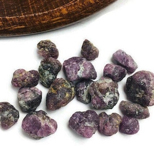 May include: A collection of rough, purple and black ruby gemstones. The stones are various sizes and shapes, with some having a more rounded appearance and others being more angular.