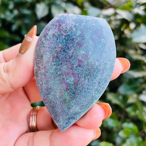 Puede incluir: Una piedra pulida en forma de lágrima con una superficie moteada. La piedra presenta una mezcla de colores, incluyendo azul profundo, verde y parches de rojo y púrpura. La piedra se sostiene en una mano, con un fondo verde borroso.
