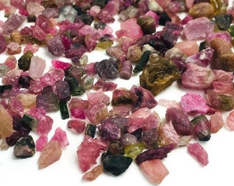 May include: A collection of pink, green, and black tourmaline gemstones. The rough, uncut stones are scattered on a white surface.