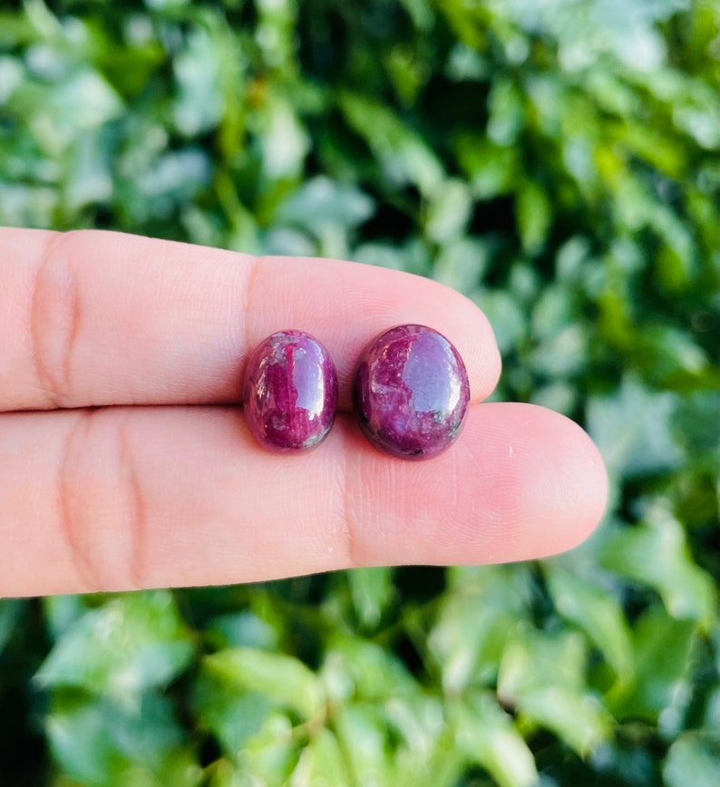 May include: Two round, cabochon-cut ruby gemstones. The stones are a deep red color with a slight purple hue. They are both about the same size and have a smooth, polished finish.