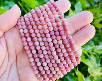 May include: A strand of pink and white gemstone beads, likely made of rhodonite, is held in a hand. The beads are round and smooth, and the strand is long enough to wrap around a wrist multiple times.