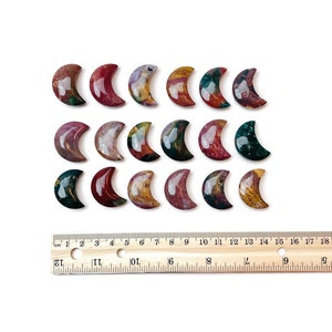 May include: A collection of 18 polished agate crescent moon shaped stones. The stones are a mix of red, brown, green, and white colors. The stones are arranged in rows on a white surface with a wooden ruler for scale.