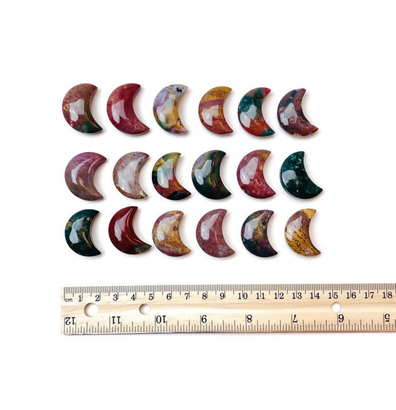 May include: A collection of 18 polished agate crescent moon shaped stones. The stones are a mix of red, brown, green, and white colors. The stones are arranged in rows on a white surface with a wooden ruler for scale.