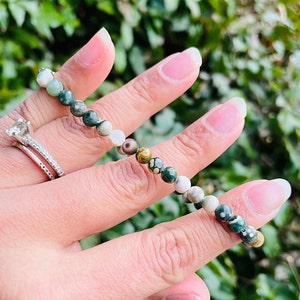 May include: A green, white, and brown gemstone bracelet with small round beads. The bracelet is on a person's wrist.