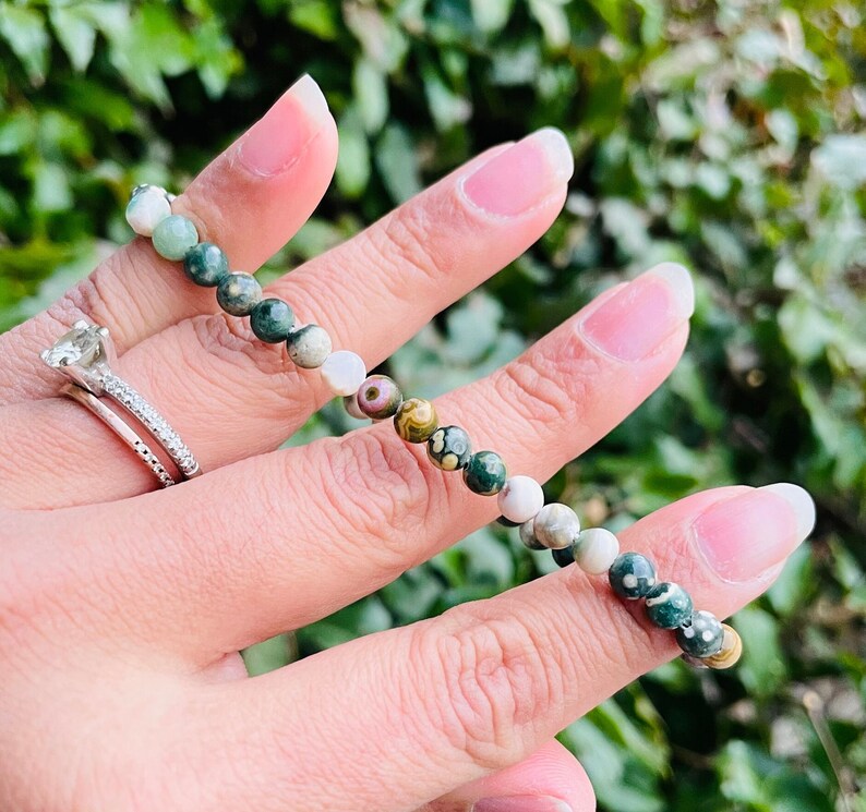 May include: A green, white, and brown gemstone bracelet with small round beads. The bracelet is on a person's wrist.