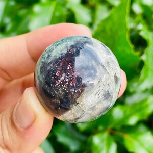 May include: A clear, spherical crystal ball with internal inclusions of shimmering red, purple, and gold. The surface has a mottled appearance with areas of green and gray. The crystal is held against a backdrop of green foliage.