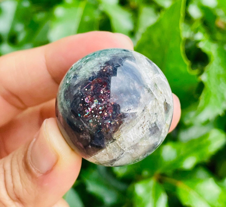 May include: A clear, spherical crystal ball with internal inclusions of shimmering red, purple, and gold. The surface has a mottled appearance with areas of green and gray. The crystal is held against a backdrop of green foliage.