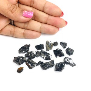 May include: A collection of small, black, rough, and irregularly shaped crystals. The crystals are scattered on a white surface.