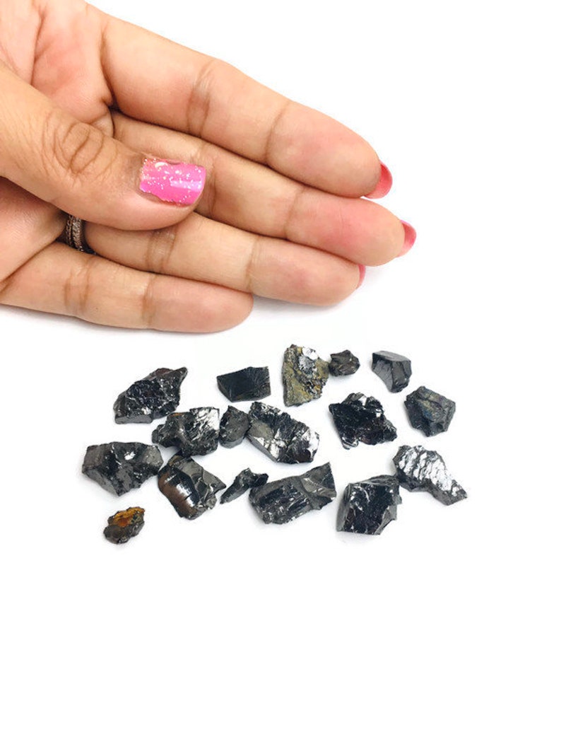 May include: A collection of small, black, rough, and irregularly shaped crystals. The crystals are scattered on a white surface.