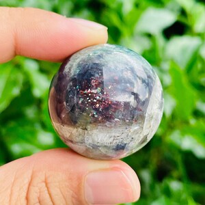 May include: A clear, iridescent sphere with a mix of colors, including purple, green, and red, is held between fingers. The sphere has a glittery, speckled appearance. The background is blurred green foliage.