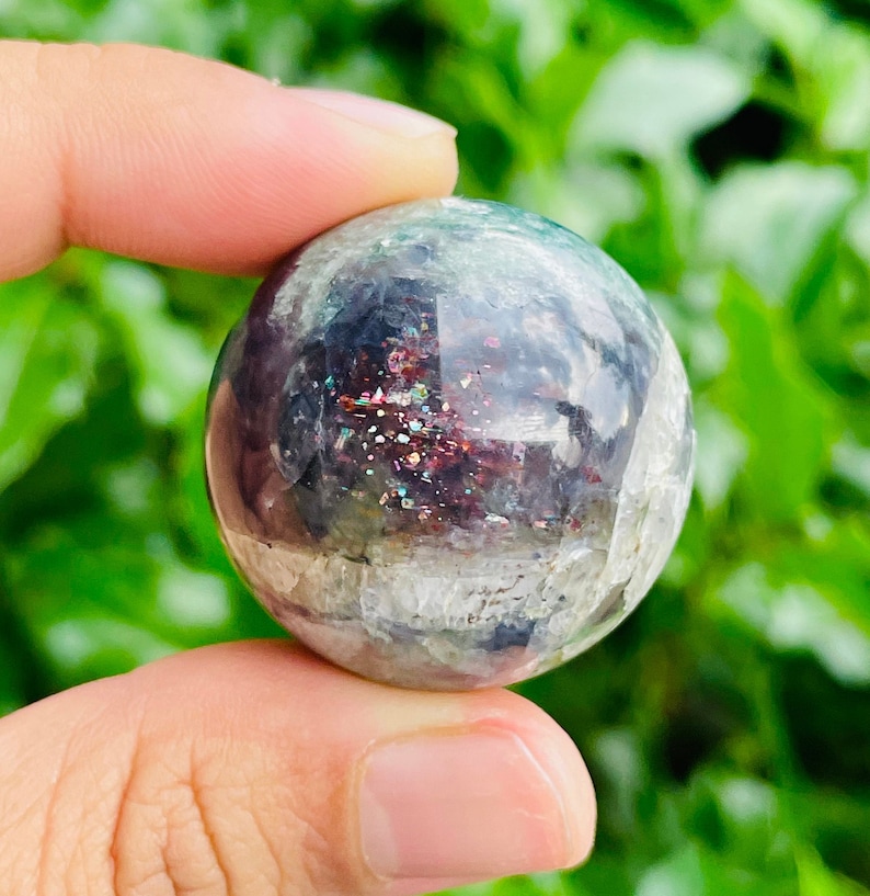 May include: A clear, iridescent sphere with a mix of colors, including purple, green, and red, is held between fingers. The sphere has a glittery, speckled appearance. The background is blurred green foliage.