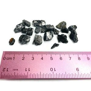 May include: A close-up of a pile of black, irregularly shaped, shiny stones on a white surface. A pink ruler with black markings is below the stones. The ruler measures 10 centimeters.