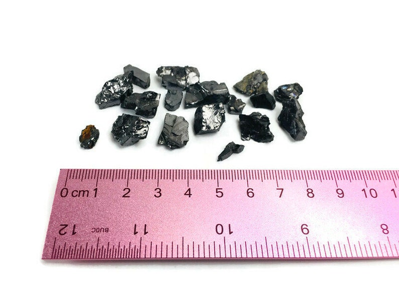 May include: A close-up of a pile of black, irregularly shaped, shiny stones on a white surface. A pink ruler with black markings is below the stones. The ruler measures 10 centimeters.
