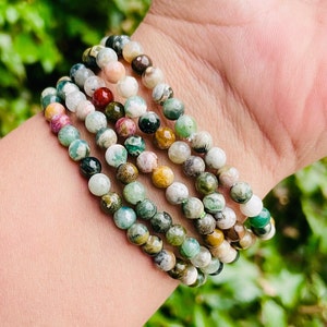 May include: A stack of five bracelets made with green, brown, and white jasper beads. The bracelets are stacked on a person's wrist.