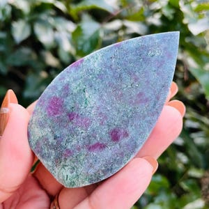 Puede incluir: Una gema pulida en forma de lágrima con un aspecto moteado. La piedra presenta una mezcla de colores, incluyendo morado intenso, verde y gris. La gema se sostiene en una mano, con un fondo verde borroso.