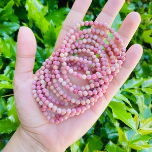 K&ouml;nnte beinhalten: Ein Stapel rosafarbener und wei&szlig;er Edelstein-Armb&auml;nder. Die Armb&auml;nder bestehen aus kleinen, runden Perlen und sind kreisf&ouml;rmig angeordnet.