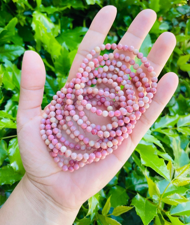 K&ouml;nnte beinhalten: Ein Stapel rosafarbener und wei&szlig;er Edelstein-Armb&auml;nder. Die Armb&auml;nder bestehen aus kleinen, runden Perlen und sind kreisf&ouml;rmig angeordnet.
