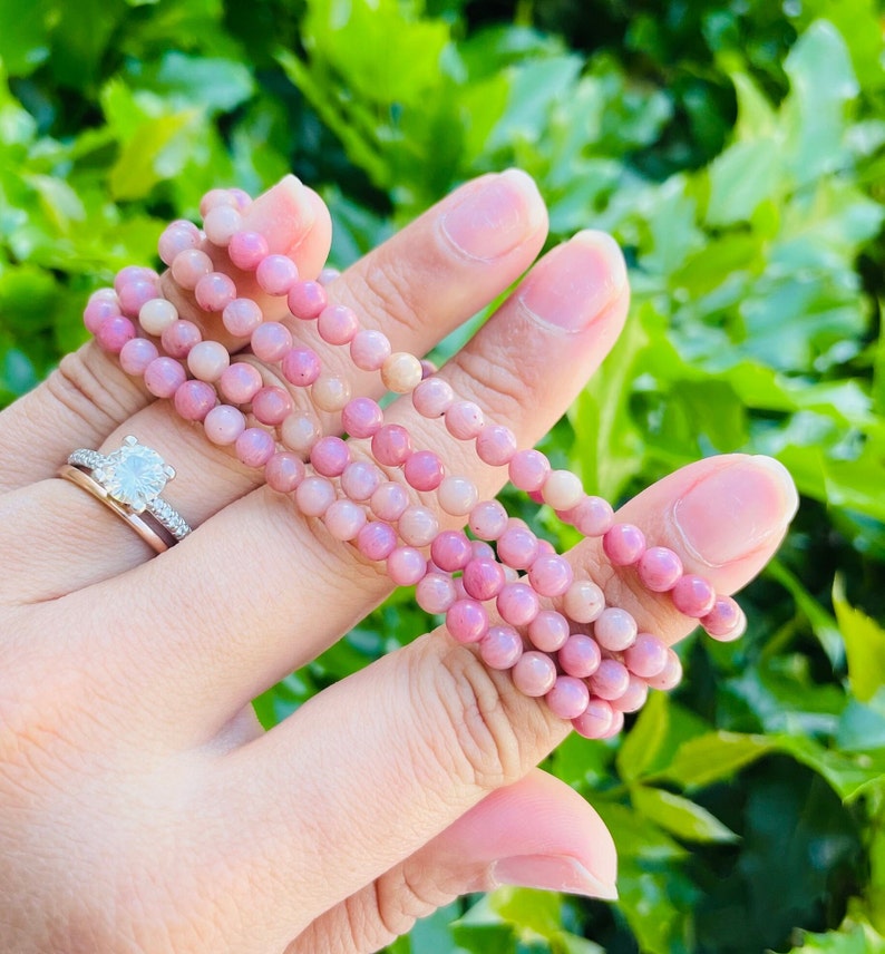 K&ouml;nnte beinhalten: Ein Stapel rosafarbener und wei&szlig;er Edelstein-Armb&auml;nder. Die Armb&auml;nder bestehen aus kleinen, runden Perlen und sind auf einem Gummiband aufgef&auml;delt.