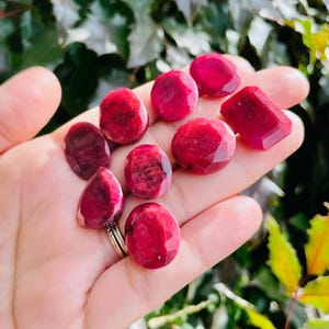 May include: A collection of eight faceted ruby gemstones in various shapes and sizes, displayed on a hand. The gemstones are a deep red color and have a polished finish.