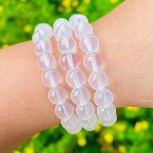 Könnte beinhalten: Drei weiße Perlenarmbänder, die auf dem Handgelenk einer Person übereinander liegen. Die Perlen sind rund und durchscheinend und wirken wie klares Glas.