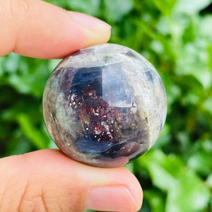 May include: A clear, spherical crystal ball with internal iridescent inclusions of red, purple, and gold. The sphere is held between fingers against a backdrop of green foliage. The crystal ball has a polished, reflective surface.