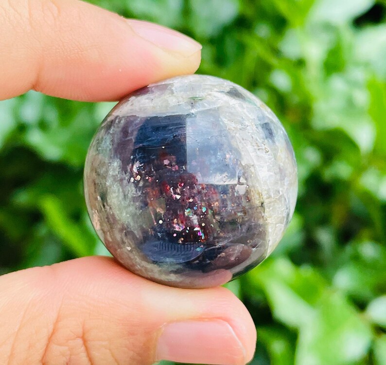 May include: A clear, spherical crystal ball with internal iridescent inclusions of red, purple, and gold. The sphere is held between fingers against a backdrop of green foliage. The crystal ball has a polished, reflective surface.