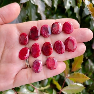 Puede incluir: Una colección de 12 piedras preciosas de rubí rojo, cada una con una forma ovalada facetada. Las piedras preciosas se muestran en una mano, mostrando su color vibrante y sus superficies pulidas.