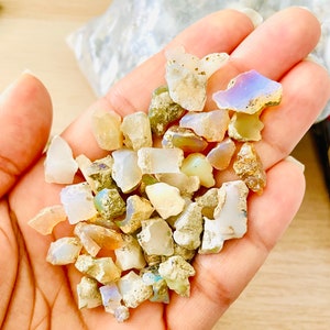 May include: A collection of small, iridescent opal chips in various shades of white, brown, and gray. The chips are scattered on a hand, showcasing their natural beauty and unique patterns.