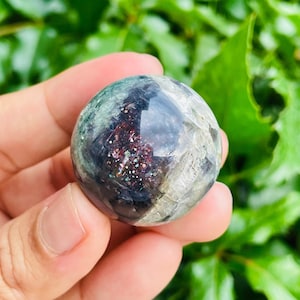 May include: A clear, spherical crystal ball with a mix of colors, including green, gray, and red. The crystal has a reflective surface and is held in a hand against a backdrop of green foliage. The crystal ball is a decorative item.