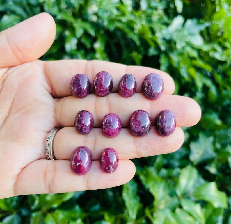 May include: A collection of ten oval-shaped, polished ruby gemstones in various shades of red and purple. The gemstones are arranged in a hand, with the largest gemstone in the center.