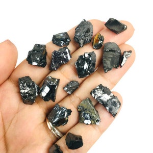 May include: A collection of black, rough, and irregularly shaped crystals. The crystals are scattered on a hand, showcasing their natural, raw form.