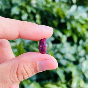 May include: A close-up of a deep purple oval cabochon gemstone held between two fingers. The stone has a slightly cloudy appearance and a smooth, polished surface.