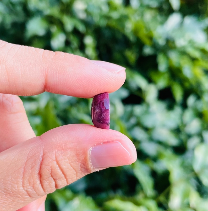May include: A close-up of a deep purple oval cabochon gemstone held between two fingers. The stone has a slightly cloudy appearance and a smooth, polished surface.