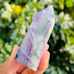 May include: A polished fluorite crystal point, showcasing a blend of purple, green, and clear hues. The crystal has a rectangular shape with a pointed top. The natural stone is held against a blurred green background.