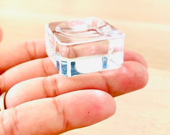 Puede incluir: Un expositor cuadrado de acrílico transparente con una plataforma central elevada. El expositor es perfecto para mostrar objetos pequeños como joyas, cristales o figuritas.