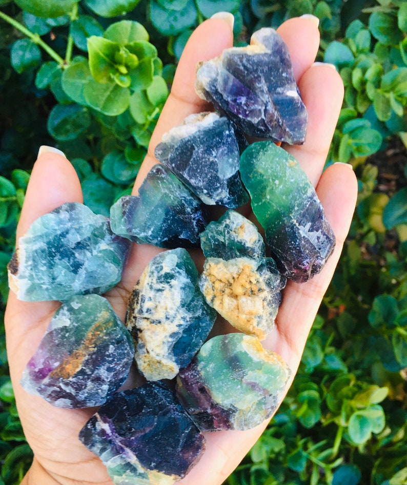 Pu&ograve; includere: Una collezione di cristalli di fluorite verde, blu e viola. I cristalli sono grezzi e hanno un aspetto naturale non lucidato.