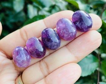 May include: Five polished amethyst stones, each with a smooth, oval shape. The stones display a range of purple hues, from light lavender to deep violet, with a glossy finish. The stones are held in a hand, with a blurred green background.