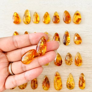 Op de afbeelding: Een verzameling amber druppel hangers, sommige met insluitsels, getoond op een licht houten oppervlak. Twee hangers worden in een hand gehouden.