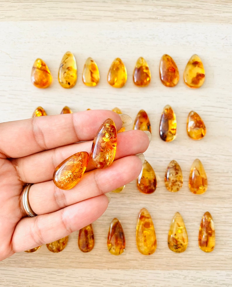 Op de afbeelding: Een verzameling amber druppel hangers, sommige met insluitsels, getoond op een licht houten oppervlak. Twee hangers worden in een hand gehouden.