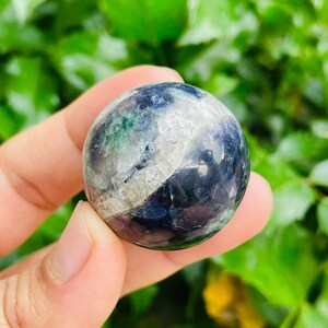 May include: A polished fluorite sphere, held in a hand, displays a mix of colors including deep blues, greens, and whites. The stone's surface has a smooth, rounded shape, with natural variations in color and texture. The background is a blurred green.