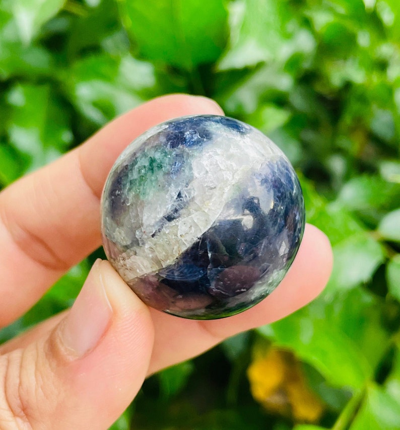 May include: A polished fluorite sphere, held in a hand, displays a mix of colors including deep blues, greens, and whites. The stone's surface has a smooth, rounded shape, with natural variations in color and texture. The background is a blurred green.
