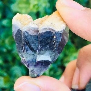 Pu&ograve; includere: Un cristallo viola, verde e bianco con una texture ruvida e naturale. Il cristallo ha la forma di un cuore ed &egrave; tenuto in una mano.