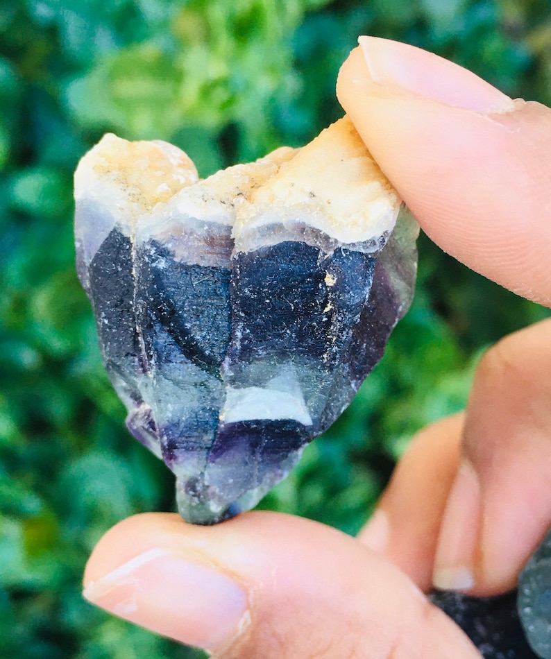 Pu&ograve; includere: Un cristallo viola, verde e bianco con una texture ruvida e naturale. Il cristallo ha la forma di un cuore ed &egrave; tenuto in una mano.