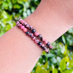 May include: Two bracelets made of pink and black gemstone beads. The bracelets are stacked on a person's wrist.